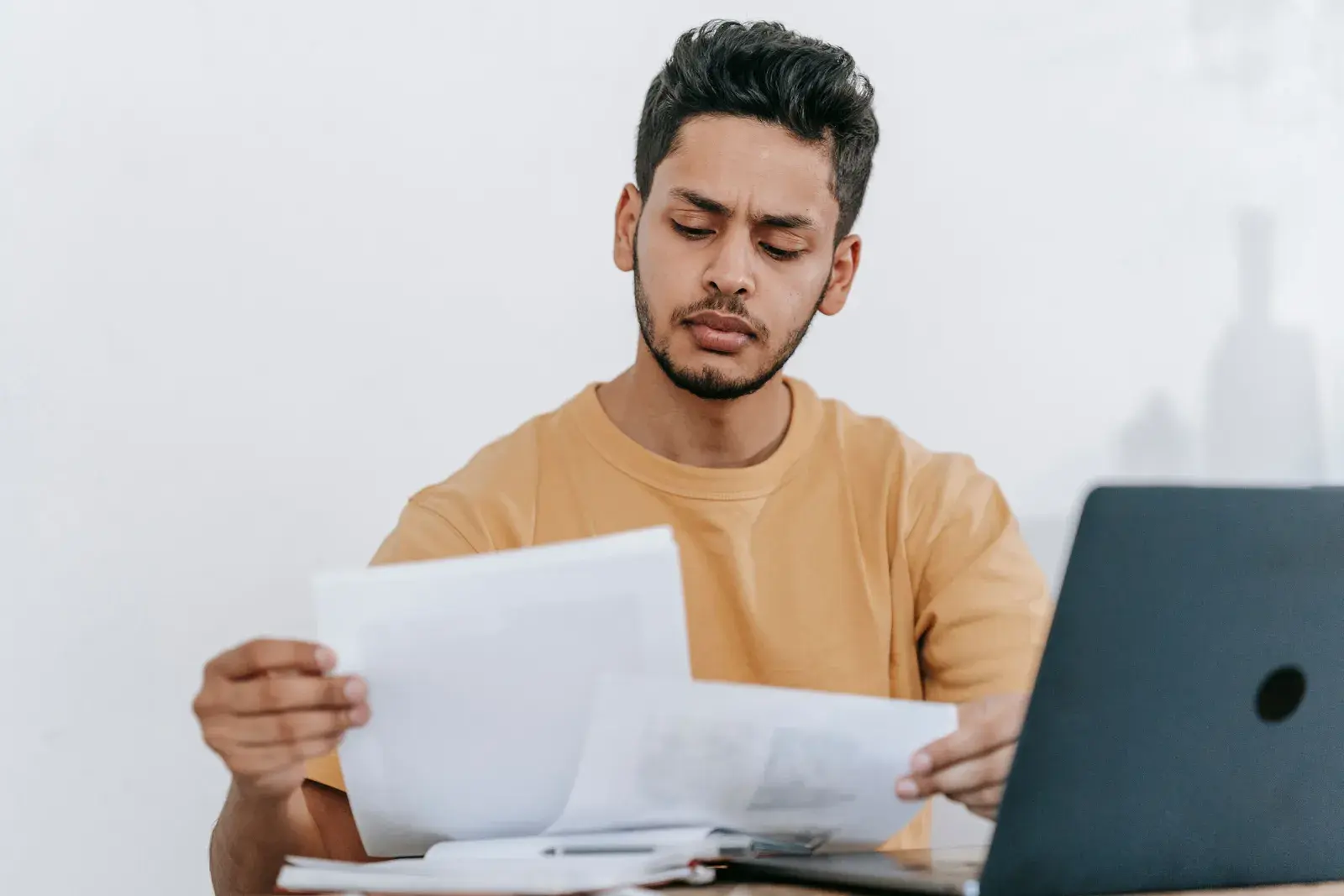 A man studying printed papers he holds in both hands beside an open laptop