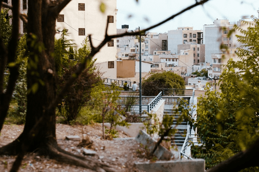 Elevated view of a dense Iranian cityscape framed by trees and a hillside walkway